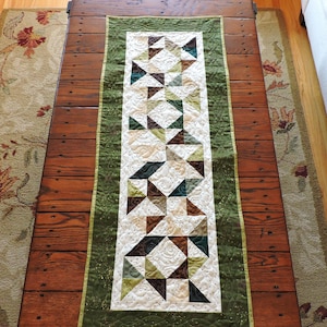 May include: A quilted table runner with a geometric pattern of triangles in shades of green, brown, and cream. The runner has a green border and is placed on a wooden table. The design is a mix of modern and traditional quilting.
