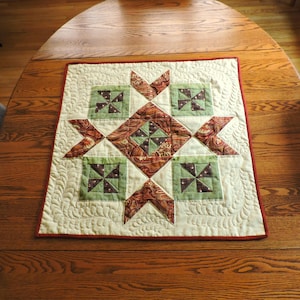 Op de afbeelding: Een vierkante stoffen quilt met een centraal ruitpatroon omringd door vier kleinere vierkante blokken met windmolenontwerpen. De quilt is gemaakt van crème, saliegroene en roestkleurige paisley-stof en afgewerkt met een donkerrode rand.