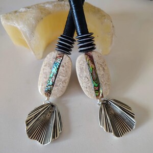 May include: A pair of handcrafted earrings featuring a combination of materials. The earrings have black cylindrical tops, textured beige oval stones with iridescent accents, and silver-colored shell-shaped pendants. The design is artistic and unique.