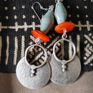 May include: A pair of handcrafted earrings featuring silver-toned, hammered-metal discs with a circular design. The earrings also include orange and blue-green beads, and silver-colored ear wires. The earrings are displayed on a patterned fabric.