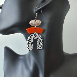 May include: A pair of dangle earrings with a black and white patterned arch, a bright orange curved piece, and a silver oval charm.