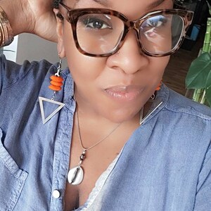 May include: A close-up of a person wearing tortoise shell glasses and a denim shirt. The person is wearing orange earrings with a silver triangle and a shell pendant necklace. The person has curly dark hair.