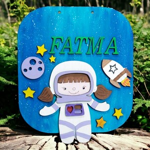 Op de afbeelding: Een blauw houten bord met de naam "FATMA" in groene letters. Het bord toont een cartoon-astronaut, een raket, sterren en een planeet. De achtergrond is blauw met witte spikkels.
