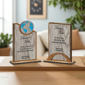 May include: Two wooden desk awards with a light wood grain finish. One features a blue globe design, the other a curved base. Both have the words "Thank You" and space for a name, ideal for recognition.
