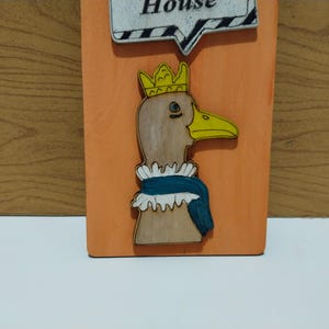 Może przedstawiać: Drewniana tablica z kresk&oacute;wkową kaczką w koronie i falbankowym kołnierzu. Kaczka ma ż&oacute;łty dzi&oacute;b i jest umieszczona na pomarańczowym tle. Chmurka z napisem "House" znajduje się na g&oacute;rze.