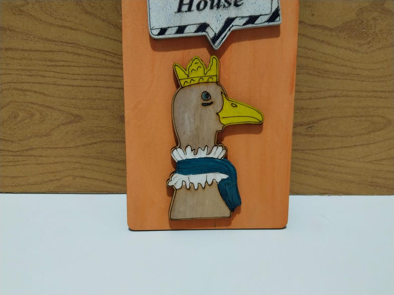 Może przedstawiać: Drewniana tablica z kresk&oacute;wkową kaczką w koronie i falbankowym kołnierzu. Kaczka ma ż&oacute;łty dzi&oacute;b i jest umieszczona na pomarańczowym tle. Chmurka z napisem "House" znajduje się na g&oacute;rze.