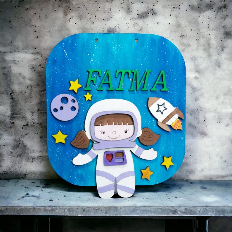Op de afbeelding: Een houten bord met een blauwe achtergrond en de naam "FATMA" in groene letters. Het bord toont een cartoon-astronaut, een raket, een planeet en gele sterren. De astronaut draagt een wit ruimtepak met paarse accenten.