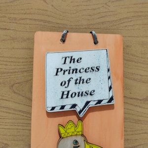 Może przedstawiać: Pomarańczowa drewniana tablica z napisem "The Princess of the House" w dymku. Na dole ilustracja księżniczki z kresk&oacute;wki. Dwa metalowe k&oacute;łka u g&oacute;ry do zawieszenia. Dekoracja ścienna.