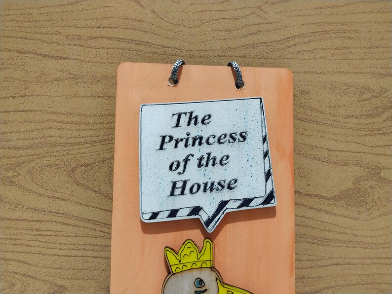 Może przedstawiać: Pomarańczowa drewniana tablica z napisem "The Princess of the House" w dymku. Na dole ilustracja księżniczki z kresk&oacute;wki. Dwa metalowe k&oacute;łka u g&oacute;ry do zawieszenia. Dekoracja ścienna.