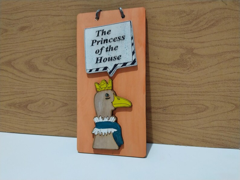 Może przedstawiać: Drewniana tablica z kaczką w koronie i niebieskim szaliku. Tablica zawiera tekst "The Princess of the House" w dymku. Tło jest w kolorze jasnopomarańczowym.