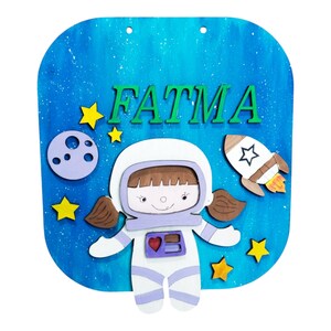 Op de afbeelding: Een houten bord met een blauwe achtergrond met een cartoon astronaut, sterren, een maan en een raket. De naam "FATMA" staat in groene letters bovenaan. Het bord is ontworpen met een ruimtethema.