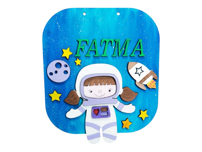 Op de afbeelding: Een houten bord met een blauwe achtergrond met een cartoon astronaut, sterren, een maan en een raket. De naam "FATMA" staat in groene letters bovenaan. Het bord is ontworpen met een ruimtethema.