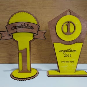 Może przedstawiać: Dwa drewniane trofea z żółtymi i brązowymi akcentami. Trofeum po lewej stronie ma baner z napisem "gratulacje" i cyfrą "1". Trofeum po prawej stronie ma cyfrę "1" w okręgu i tekst "gratulacje 2025".