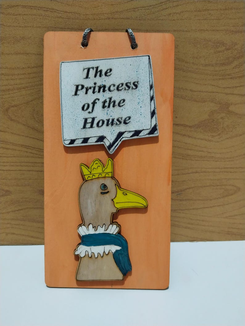 Może przedstawiać: Pomarańczowa drewniana tablica z napisem "The Princess of the House" i rysunkiem kaczki z koroną i szalikiem. Dwa metalowe uchwyty do zawieszenia.