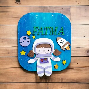Op de afbeelding: Een houten bord met een blauwe achtergrond met de naam "FATMA" in groene letters. Het bord bevat een cartoon-astronaut, een raket, sterren en een planeet. Het bord is gemonteerd op een houten oppervlak.