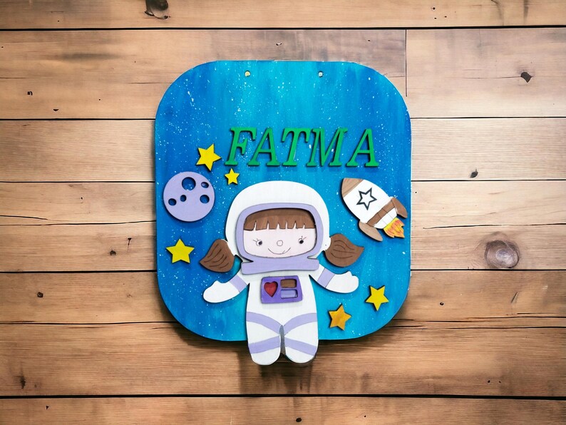 Op de afbeelding: Een houten bord met een blauwe achtergrond met de naam "FATMA" in groene letters. Het bord bevat een cartoon-astronaut, een raket, sterren en een planeet. Het bord is gemonteerd op een houten oppervlak.