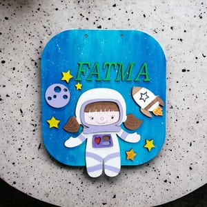 Op de afbeelding: Een blauw houten bord met de naam "FATMA" in groene letters. Het bord toont een cartoon-astronaut, een raket, een planeet en gele sterren tegen een blauw-witte achtergrond. De astronaut draagt een wit ruimtepak met paarse accenten.