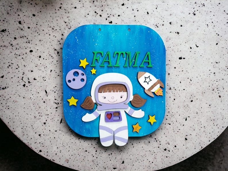 Op de afbeelding: Een blauw houten bord met de naam "FATMA" in groene letters. Het bord toont een cartoon-astronaut, een raket, een planeet en gele sterren tegen een blauw-witte achtergrond. De astronaut draagt een wit ruimtepak met paarse accenten.