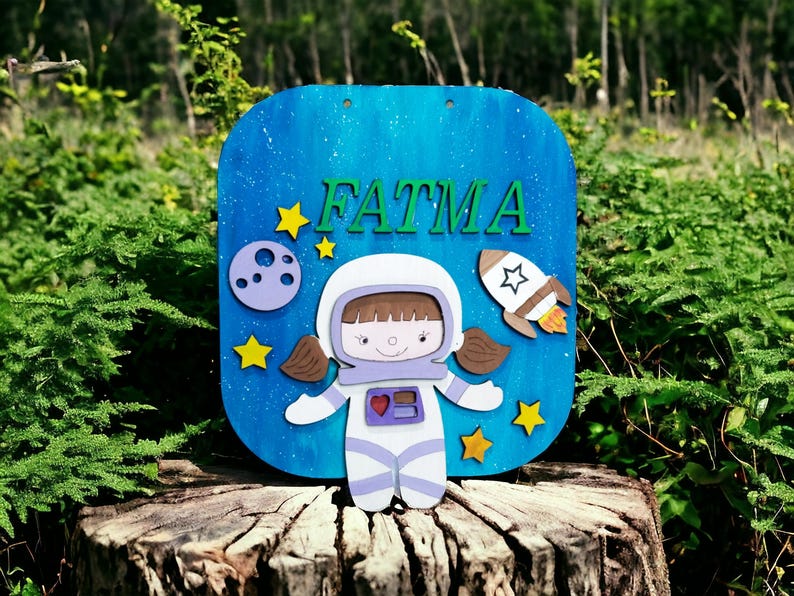 Op de afbeelding: Een blauw houten bord met de naam "FATMA" in groene letters. Het bord toont een cartoon-astronaut, een raket, sterren en een planeet. De achtergrond is blauw met witte spikkels.