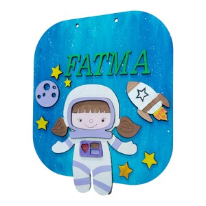 Op de afbeelding: Houten bord met een blauwe achtergrond en de naam "FATMA" in groene letters. Het bord toont een cartoon-astronaut, een raket, sterren en een maan. De astronaut draagt een wit ruimtepak met paarse accenten.