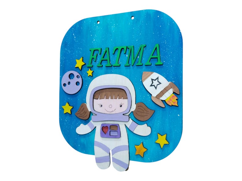 Op de afbeelding: Houten bord met een blauwe achtergrond en de naam "FATMA" in groene letters. Het bord toont een cartoon-astronaut, een raket, sterren en een maan. De astronaut draagt een wit ruimtepak met paarse accenten.