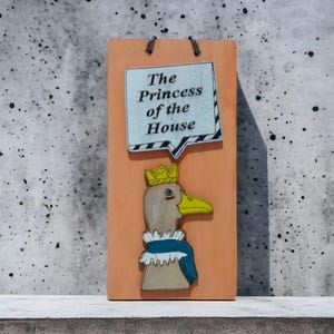 Może przedstawiać: Drewniany znak z kresk&oacute;wkową kaczką w koronie i niebiesko-białym kołnierzu. Znak zawiera tekst "The Princess of the House" w dymku. Znak jest pomarańczowy i ma metalowe pętle do zawieszania.