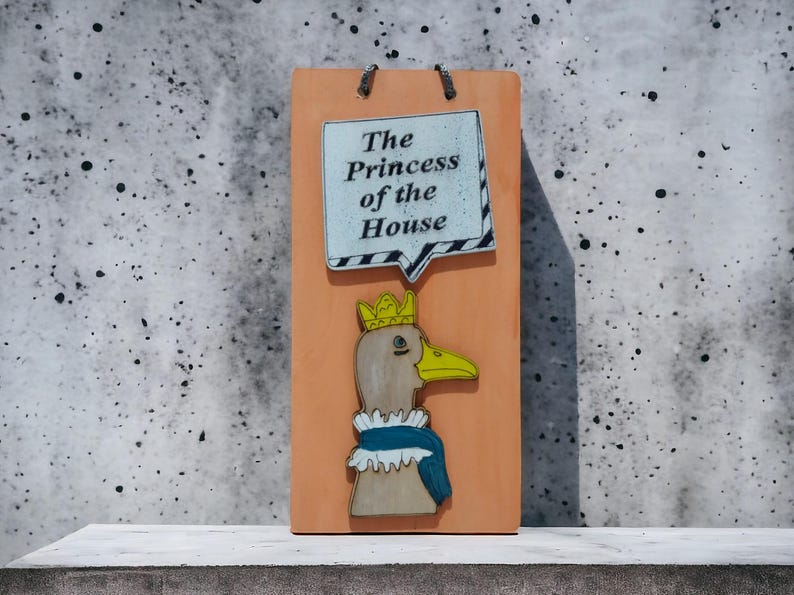 Może przedstawiać: Drewniany znak z kresk&oacute;wkową kaczką w koronie i niebiesko-białym kołnierzu. Znak zawiera tekst "The Princess of the House" w dymku. Znak jest pomarańczowy i ma metalowe pętle do zawieszania.