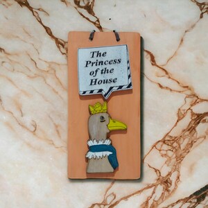 Może przedstawiać: Prostokątna drewniana tablica z kresk&oacute;wkową gęsią w koronie i niebieskim szaliku. Tablica zawiera tekst "The Princess of the House" w dymku. Tło z efektem marmuru.