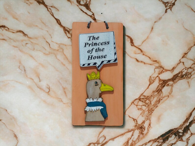 Może przedstawiać: Prostokątna drewniana tablica z kresk&oacute;wkową gęsią w koronie i niebieskim szaliku. Tablica zawiera tekst "The Princess of the House" w dymku. Tło z efektem marmuru.