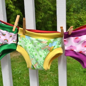 May include: Three pairs of colorful underwear hanging on a white fence. The underwear are made of cotton and have different patterns, including rainbows, hearts, and fairies.