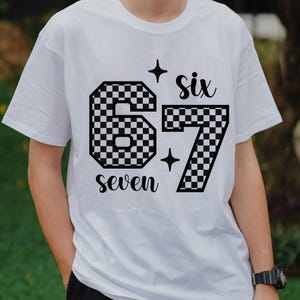 May include: White t-shirt featuring a black and white checkered design of the numbers "67" with the word "six" above and "seven" below. The design includes star accents.