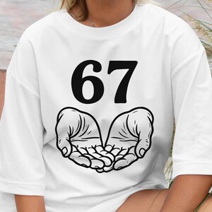 May include: White t-shirt with the number "67" in bold black font. Below the number, a graphic of two open hands cupped together is printed in black. The shirt is a casual, unisex style.
