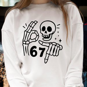 May include: White sweatshirt with a black skeleton graphic. The graphic includes a skull, a hand making the "okay" sign, and a hand pointing at the number "67". The design also features small star accents.