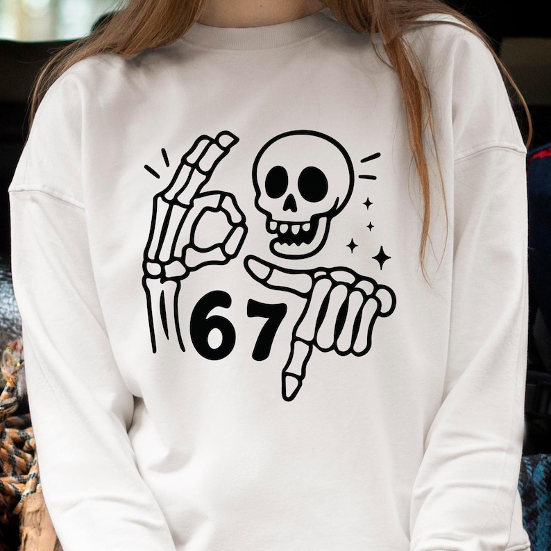 May include: White sweatshirt with a black skeleton graphic. The graphic includes a skull, a hand making the "okay" sign, and a hand pointing at the number "67". The design also features small star accents.