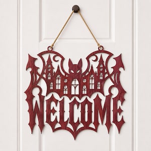 May include: A dark red, laser-cut wooden wall sign featuring a bat silhouette integrated with a gothic castle design and the word WELCOME in a sharp, pointed font. The sign is suspended by a thin, light brown jute rope from a small circular hook.
