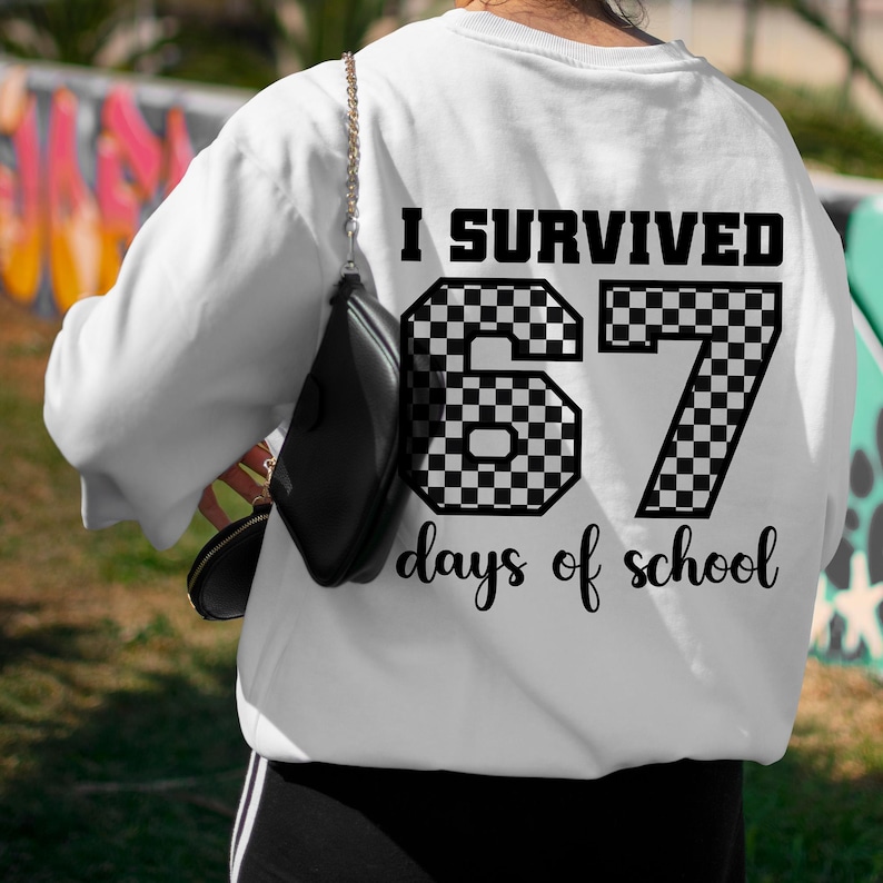 May include: White sweatshirt with the text "I SURVIVED 67 days of school" in black. The numbers are checkered. The sweatshirt is worn by a person with a black purse.