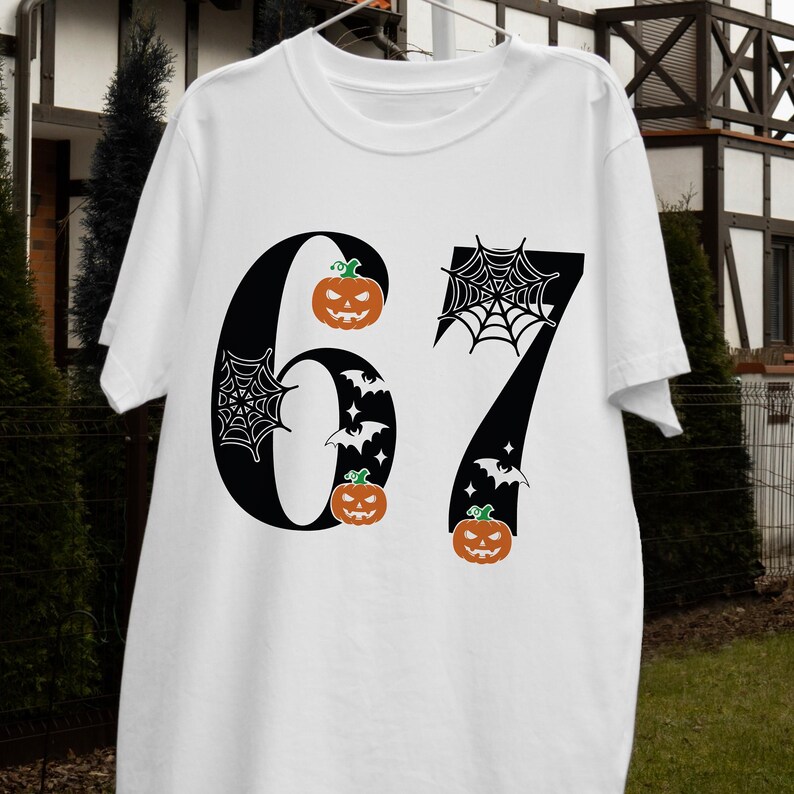 May include: White t-shirt with a black graphic of the number 67. The number 6 has a jack-o'-lantern and spiderweb design, while the 7 has a spiderweb, bats, and jack-o'-lantern design. The shirt is a Halloween-themed apparel.