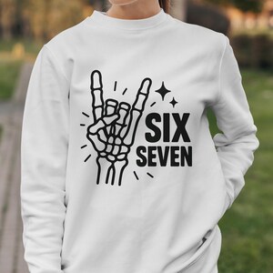 May include: White sweatshirt featuring a black skeleton hand making a rock and roll gesture. The words "SIX SEVEN" are printed in black below the hand, with star and dash accents.