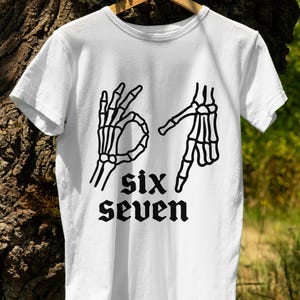 May include: White t-shirt featuring a black graphic of two skeleton hands forming the numbers six and seven in American Sign Language. The words "six" and "seven" are printed below the hands in a gothic font.