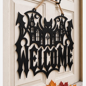 May include: A black laser-cut wooden wall hanging sign featuring a bat silhouette integrated with a gothic castle design and the word WELCOME in a sharp, pointed font. The sign is suspended by a tan rope cord from a black metal hook.