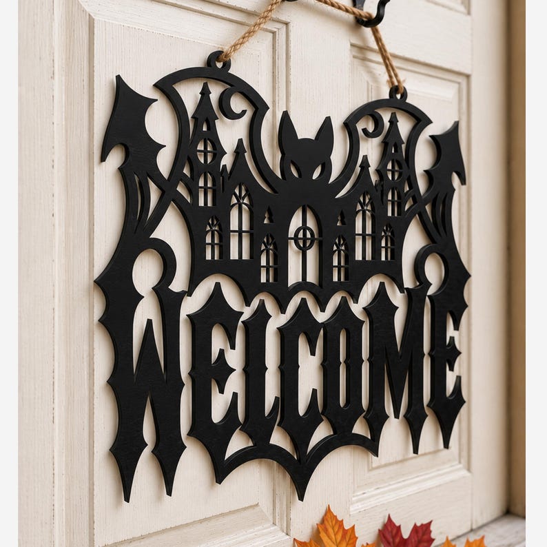 May include: A black laser-cut wooden wall hanging sign featuring a bat silhouette integrated with a gothic castle design and the word WELCOME in a sharp, pointed font. The sign is suspended by a tan rope cord from a black metal hook.