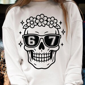 May include: White sweatshirt featuring a black and white skull design. The skull wears sunglasses with the numbers "67" and a floral crown. Four black diamond shapes and flowers surround the skull.