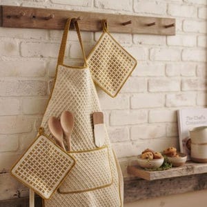 Peut inclure: Un tablier crocheté couleur crème avec une bordure jaune moutarde et une poche avant, contenant deux cuillères en bois et une spatule. Deux maniques carrées assorties au crochet sont fixées, l'une au tablier et l'autre au porte-ustensiles.