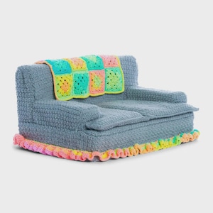 May include: A small, gray, textured sofa featuring a crocheted granny square blanket draped over the backrest. The blanket displays a vibrant pattern of neon pink, yellow, and green squares. The base of the sofa is trimmed with a ruffled edge.