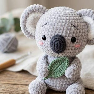 May include: A gray crocheted koala plush toy with white inner ears, black eyes, a dark gray nose, and pink cheeks. The koala is holding a small, green crocheted leaf in its hands. The texture of the yarn is soft and plush, creating a rounded shape.