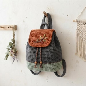May include: A crochet backpack featuring a dark gray main body, a light green base, and a rust-colored flap. The flap is decorated with an embroidered branch with green and brown leaves. Two braided drawstrings with wooden beads secure the bag.