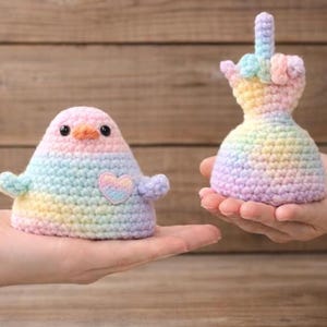 Puede incluir: Dos figuras hechas a mano con ganchillo en colores pastel arcoíris. Una es una forma de pájaro con un corazón, la otra es una mano haciendo la peineta. Ambas se sostienen en manos sobre un fondo de madera.