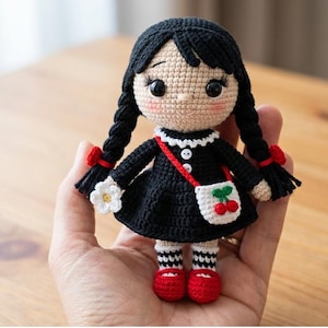 Könnte beinhalten: Gehäkelte Puppe mit schwarzen Zöpfen, trägt ein schwarzes Kleid mit weißem Kragen und eine rote Handtasche mit Kirschmuster. Die Puppe hat rote Schuhe und hält eine weiße Blume.