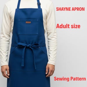 Könnte beinhalten: Eine marineblaue Schürze mit einer großen Fronttasche und einem kleinen Lederaufnäher. Die Schürze hat eine Bindung an der Taille und einen verstellbaren Nackenriemen. Der Text auf dem Bild lautet "SHAYNE APRON", "Adult size" und "Sewing Pattern".