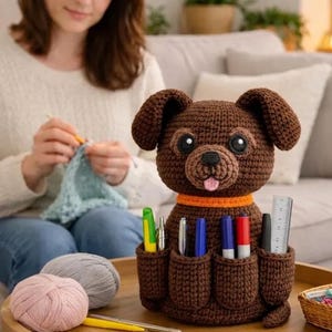 May include: A brown crocheted dog-shaped organizer with multiple pockets holding pens and a ruler. The dog has large black eyes, a pink tongue, and an orange collar. Yarn and crochet hooks are on a wooden tray in front of the dog.